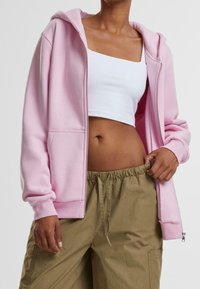 Sudadera rosa claro con cremallera, hecha de tela suave, con capucha y bolsillos delanteros, combinada con pantalones verde oliva con cordón.