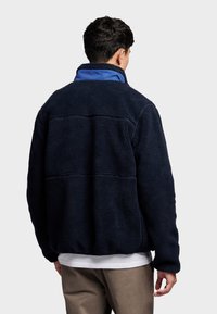 Dunkelblaue Fleecejacke mit hohem Kragen, strukturiertem Material und kontrastierender blauer Innenseite. Sie hat einen lässigen Schnitt und sichtbare Nähte.