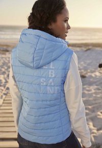 Helle blaue, gepolsterte Weste mit Kapuze, die eine horizontale Steppoptik aufweist und auf dem Rücken mit weißer Schrift versehen ist, getragen über einem langärmligen weißen Oberteil.