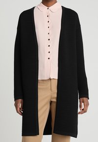 Svart ribbad cardigan med en avslappnad passform, öppen fram och långa ärmar, lagd över en ljusrosa skjorta med krage och vågiga kanter.