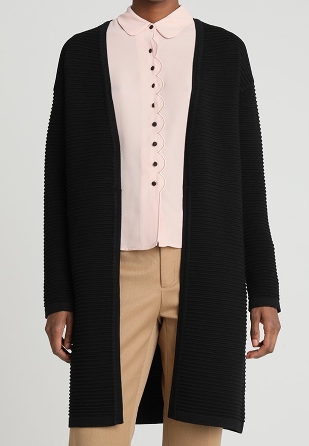 Svart ribbad cardigan med en avslappnad passform, öppen fram och långa ärmar, lagd över en ljusrosa skjorta med krage och vågiga kanter.