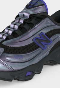 Gros plan de la basket New Balance 1000 noire et violet métallisé avec le logo "N" violet et un détail unique de protège-lacets rainuré.