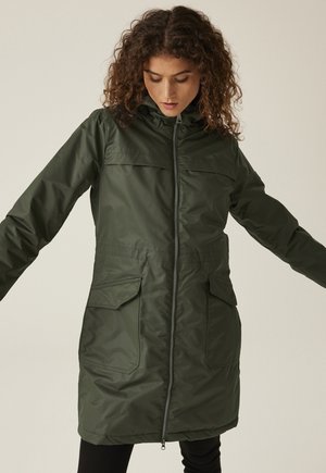 Femme portant une longue veste à capuche vert foncé avec une fermeture éclair à l'avant et de grandes poches à rabat, regardant vers le bas avec les bras légèrement étendus.