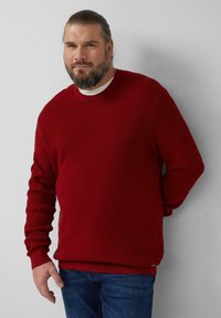 Roter, gerippter Pullover mit Rundhalsausschnitt. Das Material wirkt strukturiert. Kombiniert mit blauen Jeans, stehend vor neutralem Hintergrund.