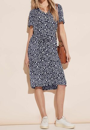 Žena ve šatech s geometrickým vzorem v námořnické modři a bílé barvě s páskem v pase, bílé tenisky a hnědá kožená kabelka přes rameno.