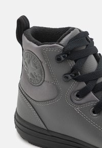 Grå hög stövel gjord av texturerat syntetmaterial. Har svarta snören, svart textilfoder och en cirkulär logotyp på utsidan.