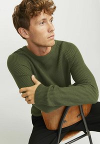 Olijfgroen geribd sweater met lange mouwen, ronde halslijn en textuurafwerking. Model zit op een houten stoel.