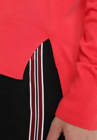 Pull en jersey corail avec fente sur le côté. Présente des poignets et un ourlet côtelés, associé à un tissu noir arborant des rayures rouges et blanches. Texture douce.