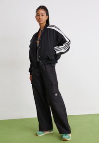 Svart bomberjakke med hvite striper, svarte cargo-bukser og sneakers med turkiske detaljer. Glatt tekstur, sporty design med fremtredende merkevare.