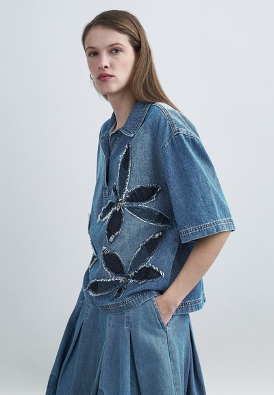 Chemise en denim avec un col, des manches courtes et une grande application fleurie en denim foncé. Associée à une jupe plissée en denim.