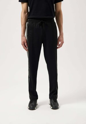 Zwarte joggingbroek met een textuurafwerking, voorzien van een trekkoord in de taille en contrasterende witte zijstrepen. G gedragen met zwarte sneakers.