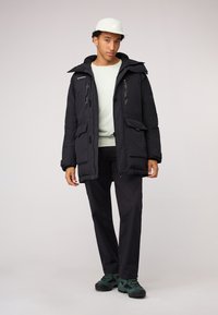 Zwarte geïsoleerde parka met capuchon, voorzien van meerdere ritszakken en een gestructureerde stof, gecombineerd met donkere broeken en groene schoenen.