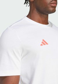 Hvid bomulds T-shirt med rund hals, korte ærmer og et lille rød-orange trekantet logo på venstre brystområde.