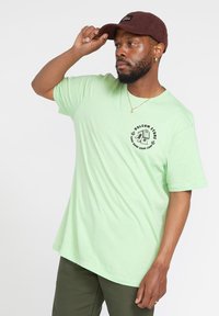 Camiseta de algodón verde claro con mangas cortas, con un logo circular negro en el pecho. Llevada con una gorra de pana en tonos tierra.