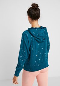 Pull-over en teal avec un motif d'étoiles blanches, capuche et poignets côtelés. Texture douce et coupe décontractée, associé à un legging rose clair.
