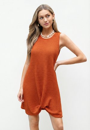 Mujer con cabello largo y rubio que lleva pendientes de aro dorados y un collar de conchas, posando con un vestido sin mangas de textura naranja y dobladillo anudado.