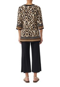 Mehrfarbige Tunika mit geometrischem Muster, Dreiviertelärmel und schwarzem Saum, kombiniert mit schwarzen weit geschnittenen Hosen und hellen Sandalen.