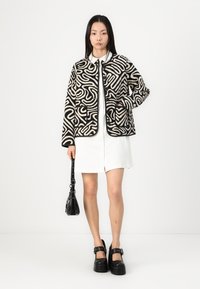 Jachetă capitonată cu un model abstract alb și negru, cu butoane în față și buzunare laterale, purtată cu o rochie albă și pantofi cu platformă negri.