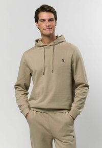 Beige kapucnis felső, húzózsinórral, apró fekete logóval. Puha anyagból készült, bordázott mandzsettákkal és laza szabással, összeillő nadrággal párosítva.
