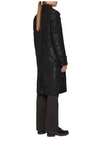 Manteau noir avec un motif floral texturé, design mi-long et col. Associé à un pantalon sombre et des chaussures noires épaisses.