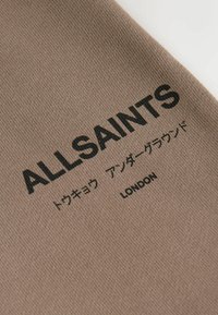 Brauner Stoff mit dem Text "ALLSAINTS" in fettgedruckten schwarzen Buchstaben, dazu weiterer Text auf Japanisch und darunter "LONDON". Strukturierte Oberfläche.
