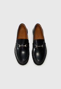 Mocasines de cuero negro con puntera redondeada, que presentan un detalle de cadena plateada, costuras texturizadas y una suela de goma de contraste.