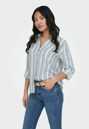 Kobieta z długimi czarnymi włosami, ubrana w niebiesko-białą pasiastą koszulę na guziki, niebieskie jeansy i beżowy pasek, pozująca z jedną ręką w kieszeni.