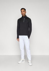 Tricou negru cu mânecă lungă și fermoar, cu guler înalt, asortat cu pantaloni albi și pantofi sport albi cu un logo închis la culoare și material texturat.