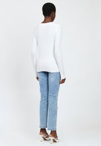 Top blanco de manga larga con textura acanalada, combinado con jeans ajustados de color azul claro y sandalias blancas de tacón, visto desde atrás.