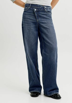 Persoon die losse donkerblauwe wijde jeans draagt met een asymmetrische geknoopte tailleband en zwarte schoenen, staand tegen een effen achtergrond.