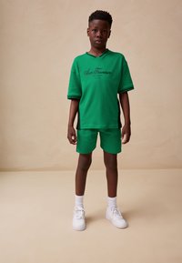 Green short-sleeve shirt and matching shorts made from soft fabric, featuring a black accent and printed logo. White sneakers complete the look.