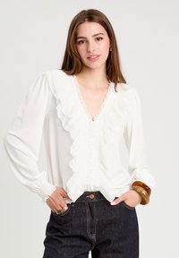 Blouse blanche avec un col et des poignets volantés, présentant un décolleté en V profond et des plis structurés. Assortie à un pantalon en denim foncé et un bracelet.