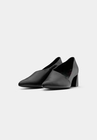 Black pointed toe heels with a smooth finish. Features a mid-height block heel and a sleek silhouette with a subtle cross-over design.
