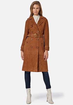 Manteau en suède marron avec une taille ceinturée, boutons à double boutonnage et col large. Porté sur un pull léger et un jean ajusté.