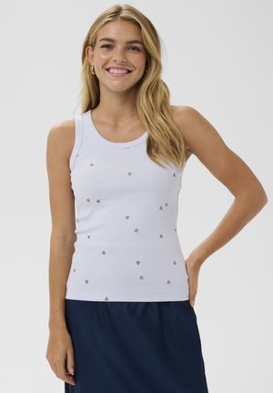 Usmievavá blondína s rozpuštenými vlasmi, oblečená v bielom bezrukávovom rebrovanom topu s malými striebornými srdiečkami a námornícky modrej sukni.