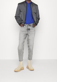 Šedá bomber bunda, královsky modré polo tričko, světle šedé džíny a béžové kotníkové boty s dřevěnou podešví. Hladké textury a strukturovaný střih.