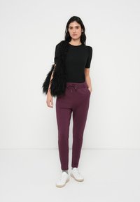 Top nero aderente, joggers bordeaux con coulisse e tasche laterali, abbinati a sneaker bianche e una borsa nera pelosa. Design semplice e moderno.