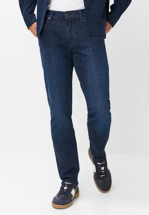 Mand iført mørkeblå jeans, navy jakke, hvid skjorte og sorte sneakers med hvide detaljer går mod en ensfarvet baggrund.