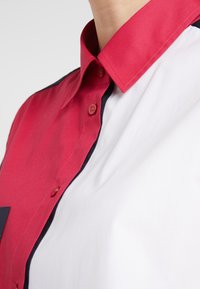 Camisa de botones roja y blanca con un ribete negro contrastante a lo largo de la botonera, con cuello estándar y textura suave.