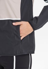 Lätt svart jacka med en beige panel, utrustad med en dragkedjeficka och elastiska ärmslut. Tillverkad av ett slätt, vattenavvisande tyg.