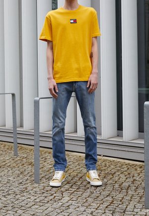 T-shirt en coton jaune avec un patch logo, associé à un jean bleu clair et des baskets avec des accents jaunes. Debout sur une surface en pierre texturée.