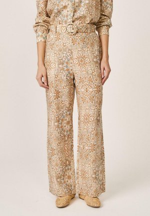 Pantalones de pernera ancha y cintura alta con cinturón a juego en tela beige con intrincados patrones florales y geométricos en tonos naranjas y marrones, combinados con zapatos beige de tejido trenzado.