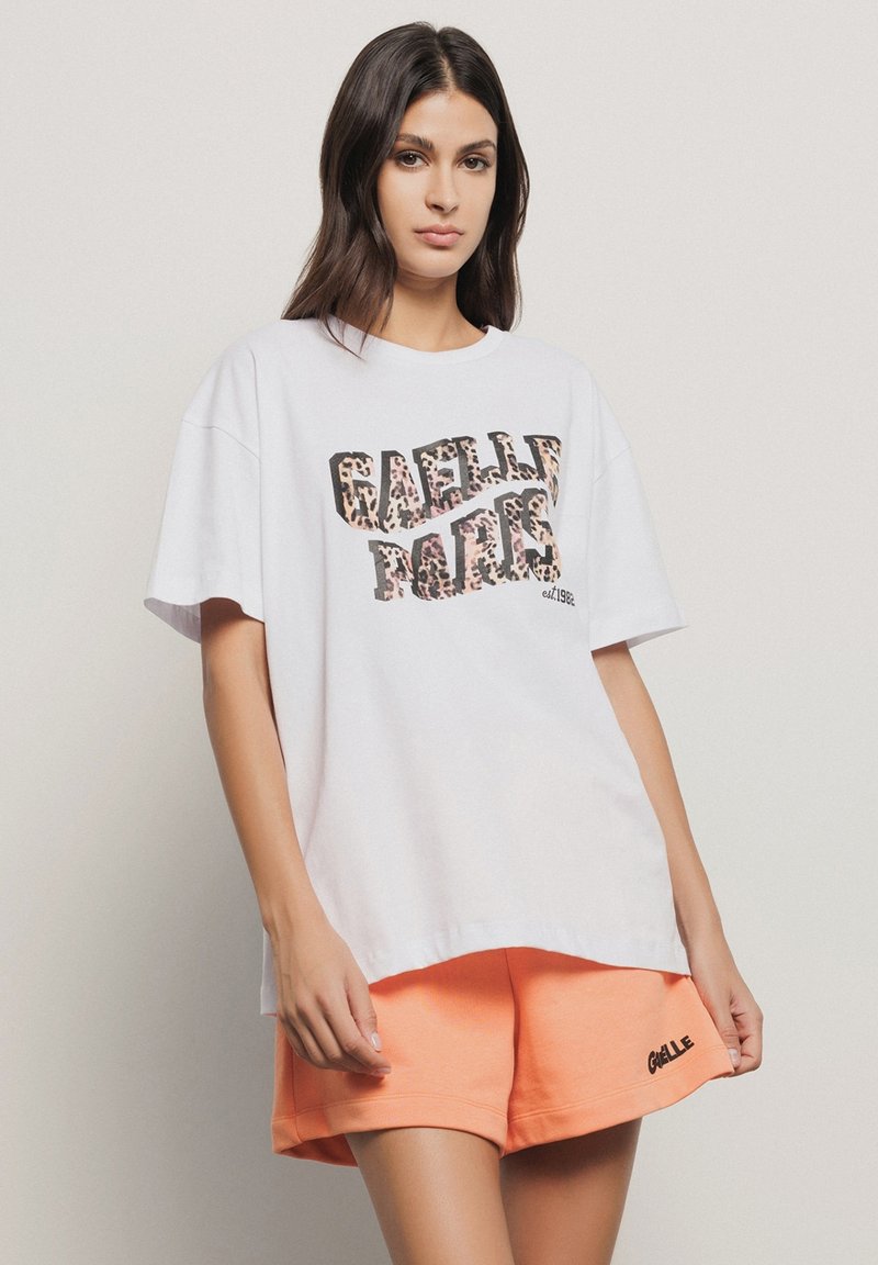 T-shirt bianco in cotone con maniche corte e testo leopardato "GAELLE PARIS" in rosa e marrone. Abbinato a pantaloni corti arancioni con logo.