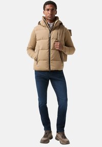 Tan puffer vest met capuchon, voorrits en zijzakken, gedragen over een beige geribde trui. Gecombineerd met blauwe jeans en een paar bruine sneakers.