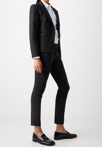 Blazer noir sur mesure avec revers en satin, chemise blanche, pantalon noir ajusté et mocassins noirs brillants. Textures lisses et coupe structurée.