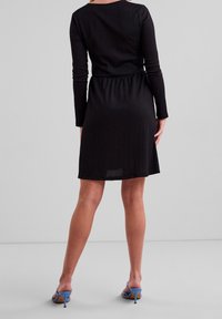 Robe noire à manches longues avec un corsage ajusté et une taille élastique, cintrant à la taille, associée à des sandales à talons bleues. Texture lisse.