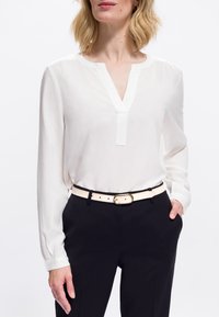 Femeie îmbrăcată cu o bluză albă cu mânecă lungă, introdusă în pantaloni negri cu o curea bej, cu mâna în buzunar și părul blond de lungime medie.