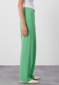 Groene, wijde broek met een textuurstof, heeft een platte tailleband, zijzakken en subtiele voorplooien. Gemaakt in combinatie met lichtgekleurde schoenen.