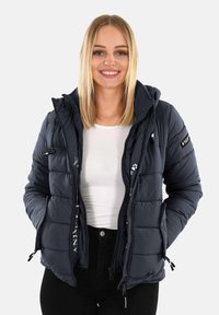 Usmievajúca sa žena s rovnými blond vlasmi má na sebe námornícku prešívanú bundu cez bielu košeľu a čierne nohavice, stojí pred jednoduchým pozadím.