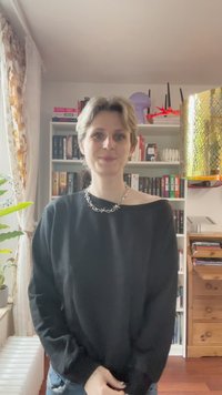 Sweater preto oversized com um design de ombro caído, colar de corrente prateada, jeans azuis, numa sala com estantes de livros e uma planta verde.